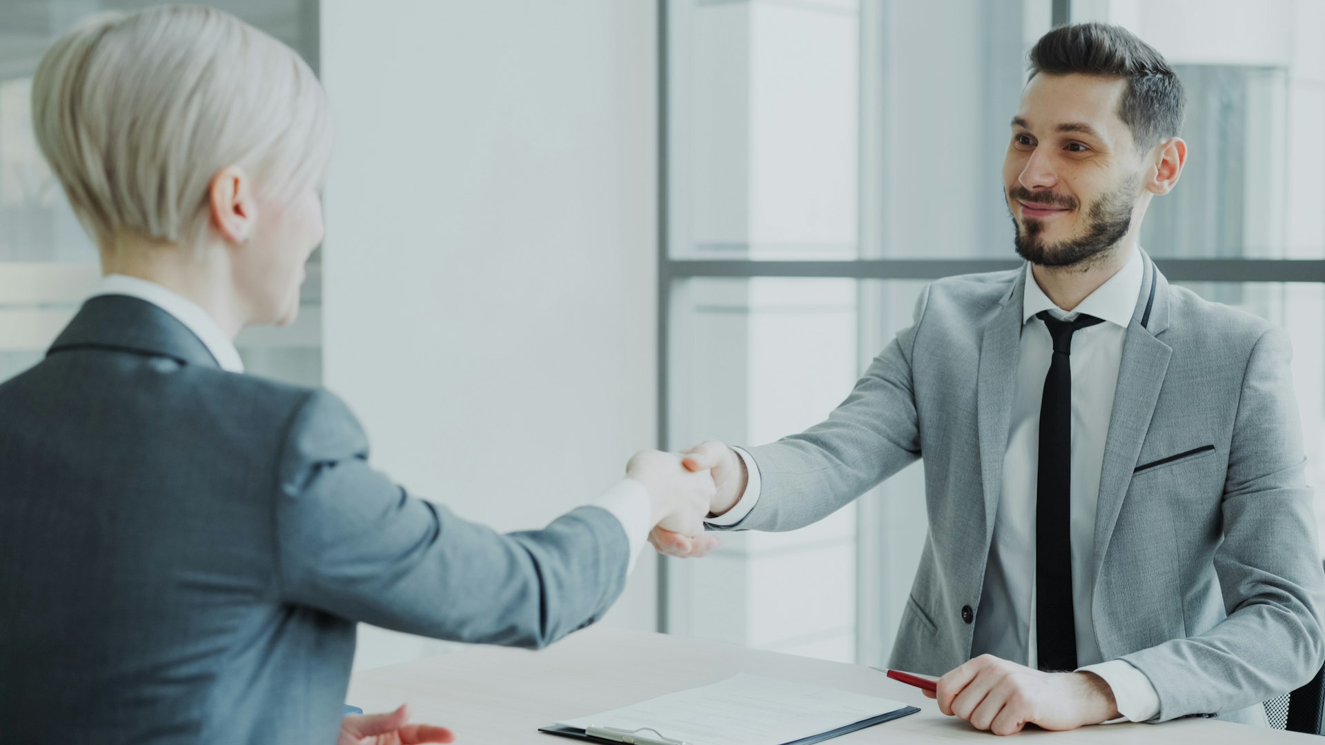 A man and a woman shaking hands across a desk in a bright office